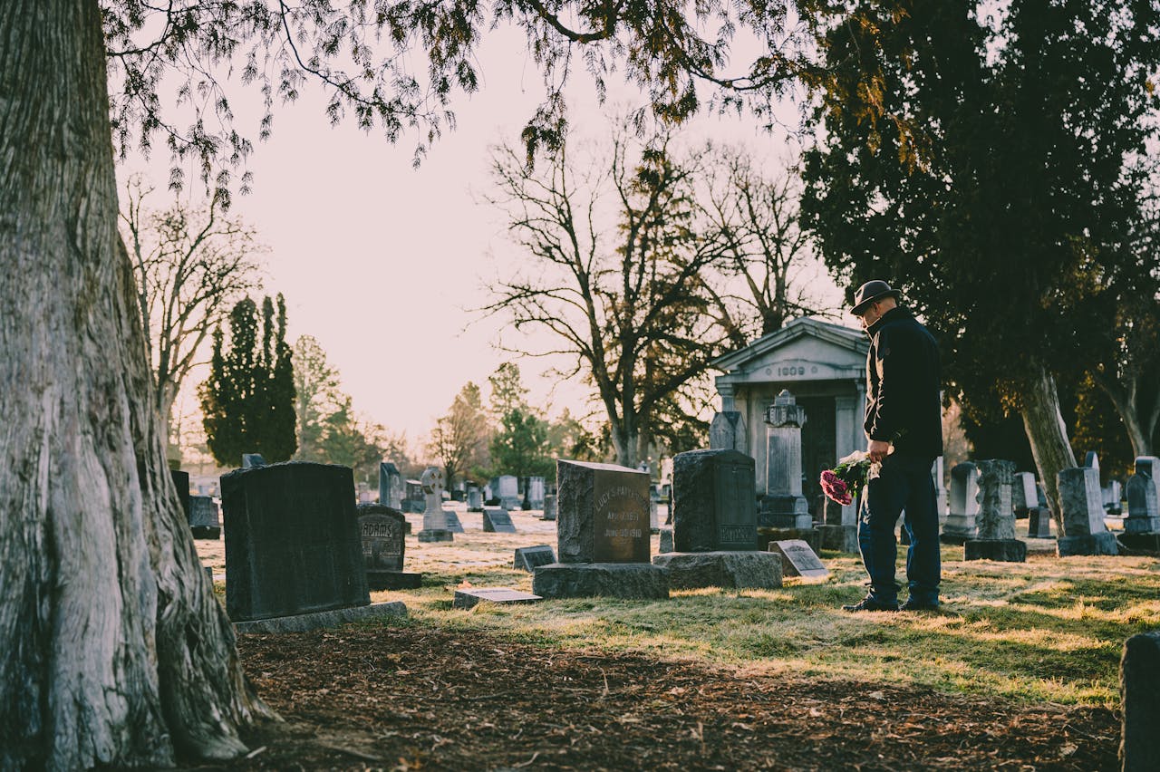 A person stands in a peaceful cemetery holding flowers in remembrance.